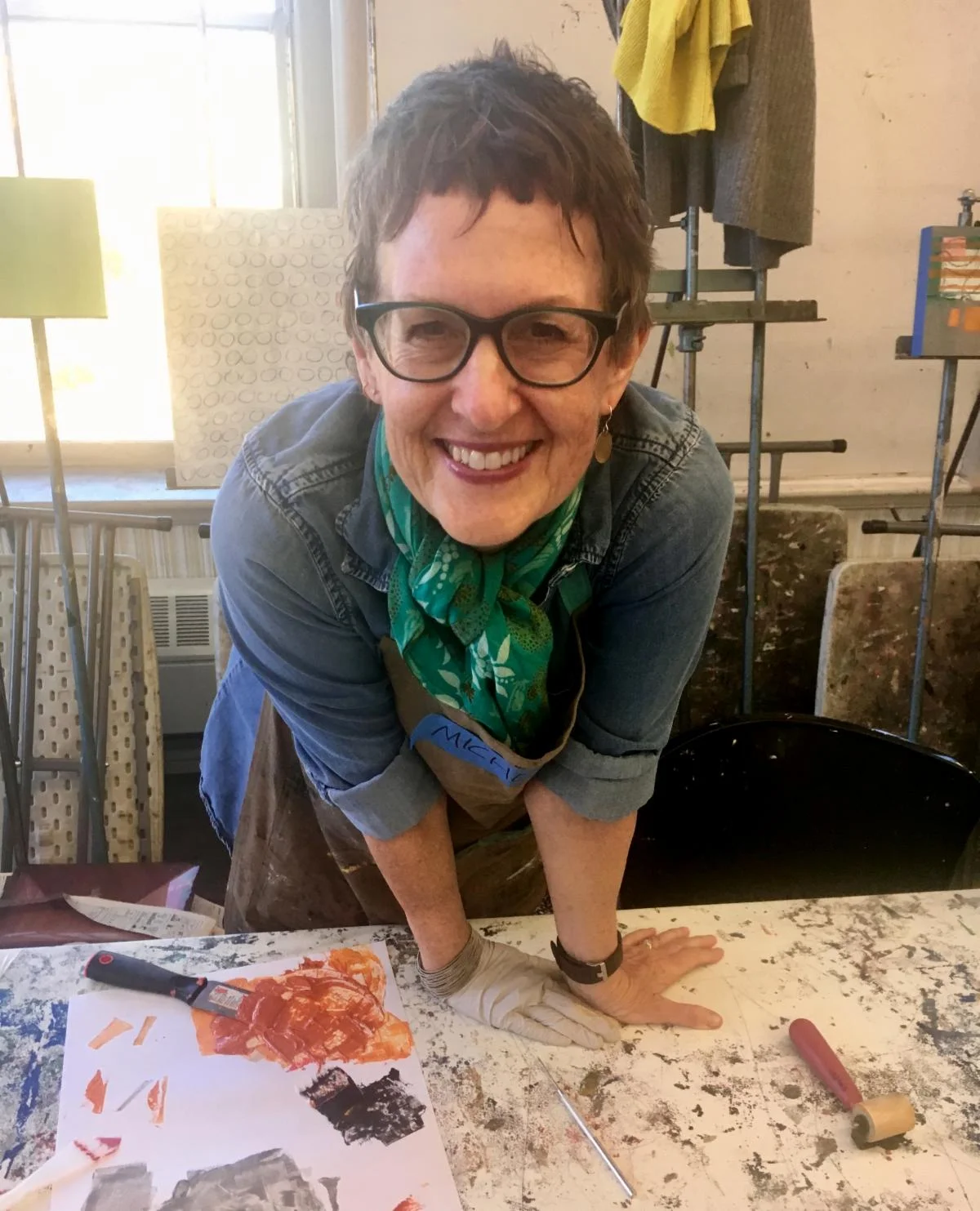 Person in denim leaning over a paint-splattered table with art supplies.